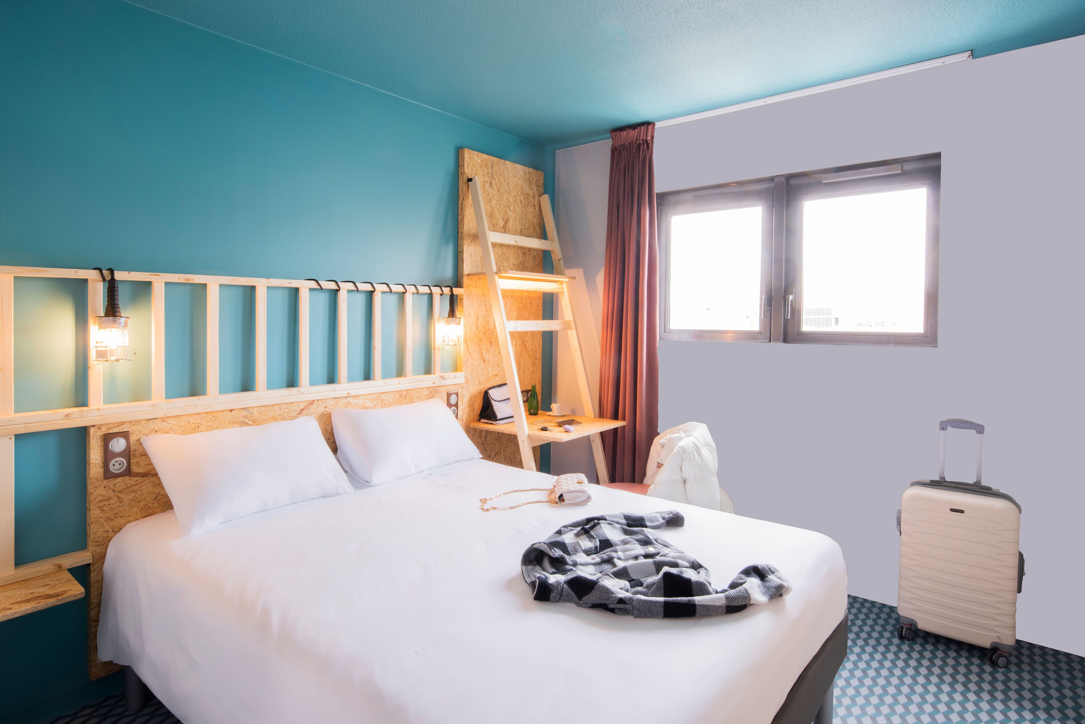 Une chambre d’hôtel avec un lit, une tête de lit en bois, un bureau avec une lampe, une valise et une fenêtre avec des rideaux.