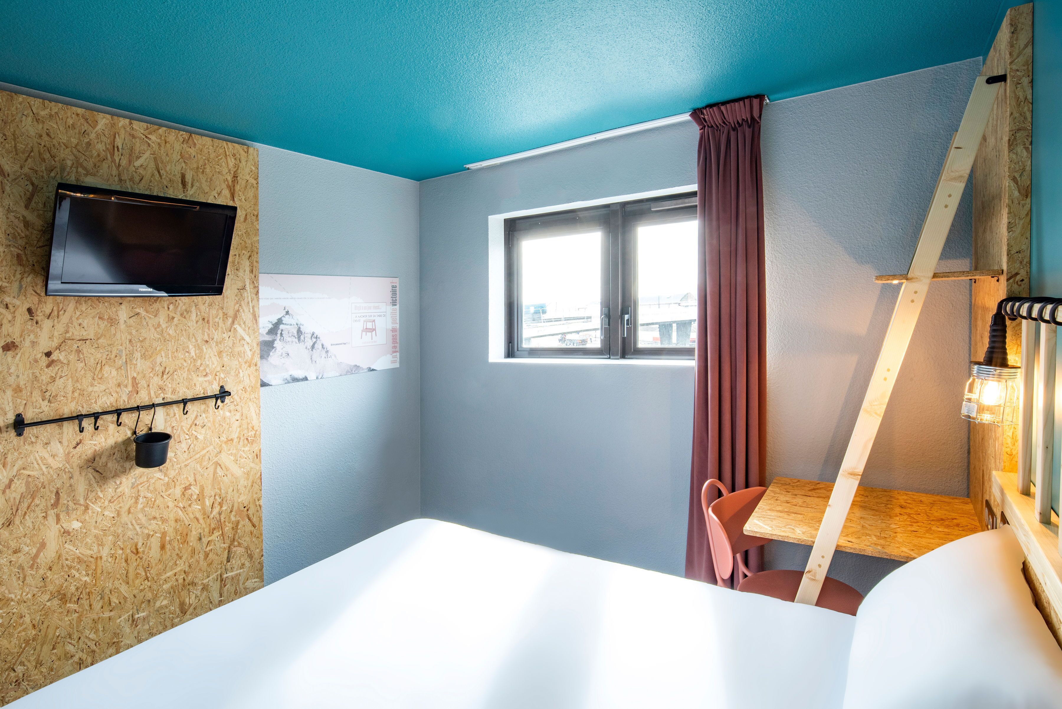 A room with a bed, a TV mounted on a wooden wall, a window with curtains, and a small table with chairs.