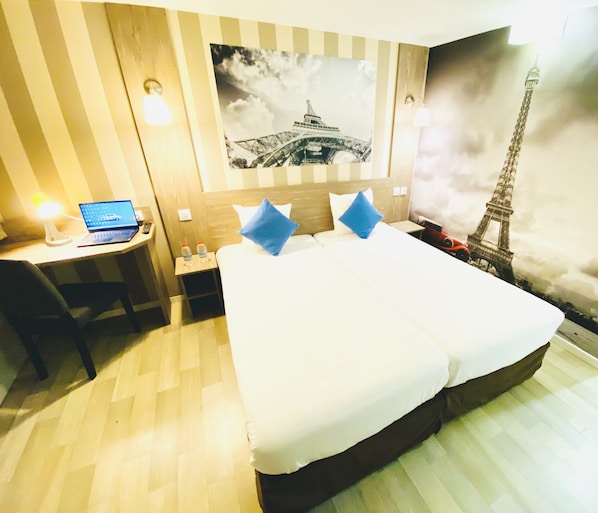 A hotel room with a bed, a desk with a laptop, a nightstand, and a wall with a picture of the Eiffel Tower.