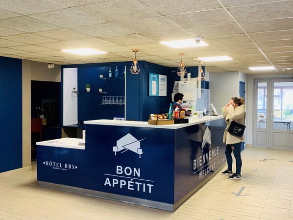 A hotel reception area with a blue counter labeled "BON APPÉTIT" and "HÔTEL RBX." There are overhead lights, a reception desk, and a person behind the counter. - Hotel RBX - Roubaix Centre (Roubaix)