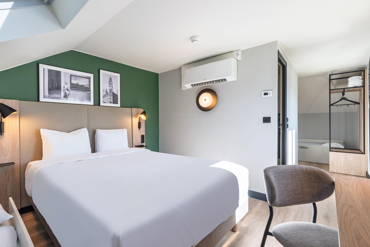 A modern hotel room with a large bed, a chair, a wall-mounted air conditioner, and framed pictures.