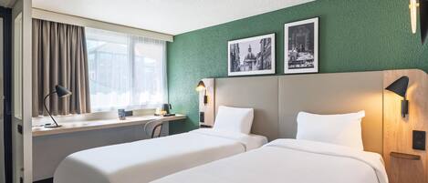 Una habitación de hotel con dos camas, un escritorio, una ventana con cortinas y dos cuadros en la pared.