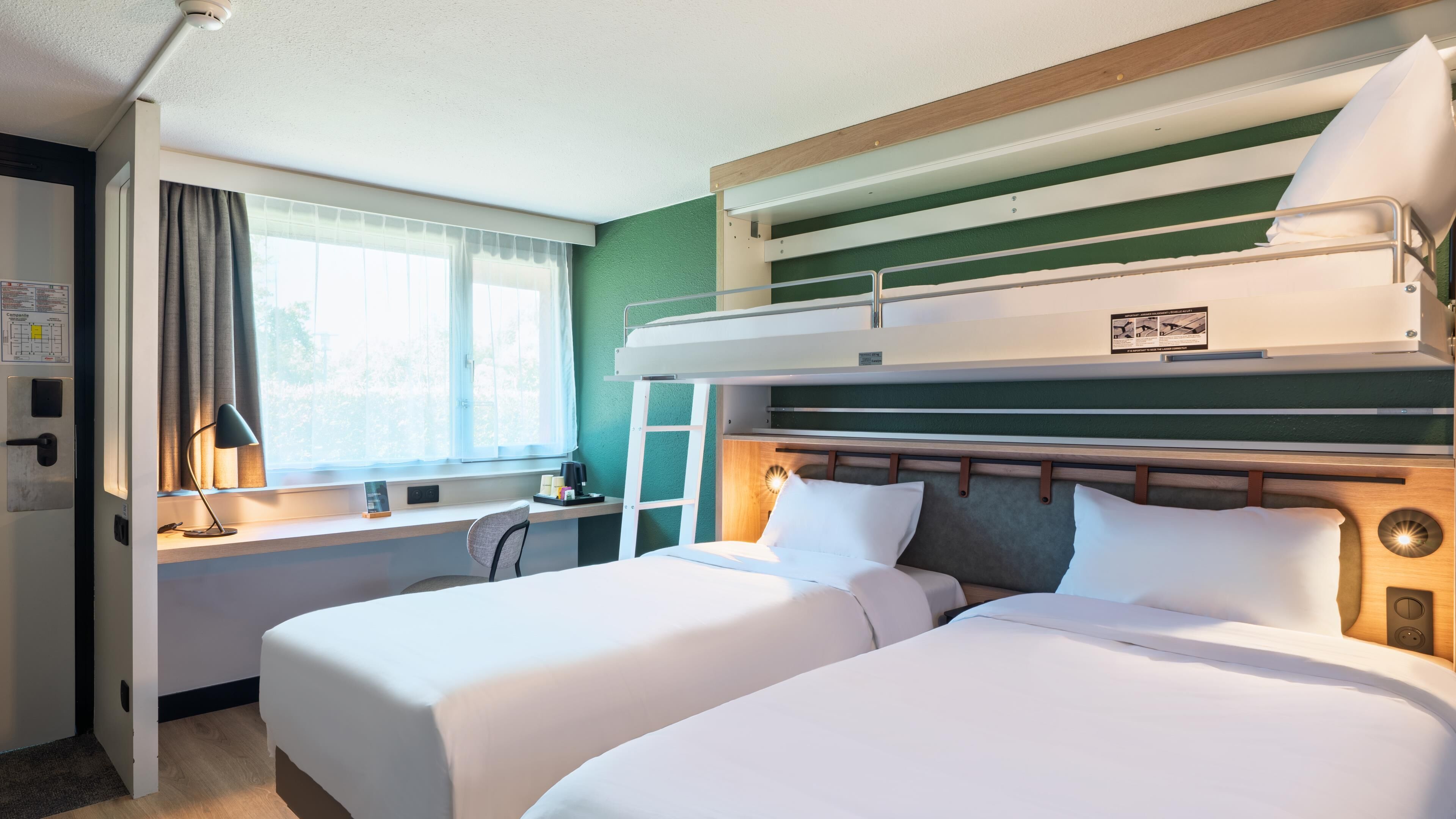 Quarto de hotel com beliches, uma escrivaninha, uma cadeira, uma janela com cortinas e uma porta.