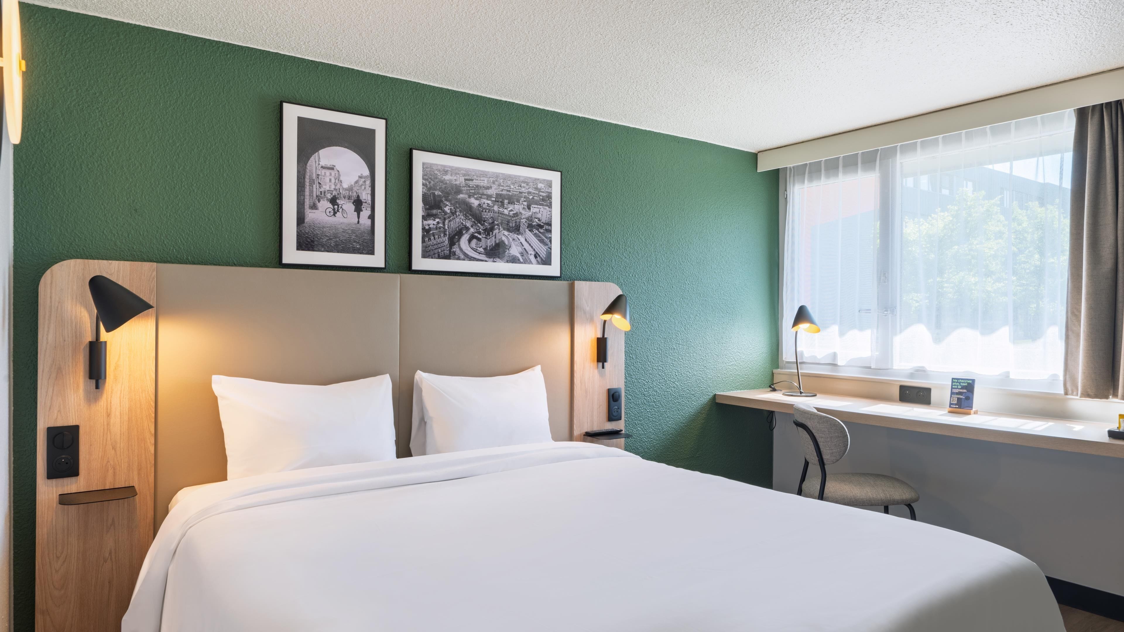 Une chambre d’hôtel avec un lit, deux photographies encadrées au mur, un bureau avec une lampe et une fenêtre avec des rideaux.