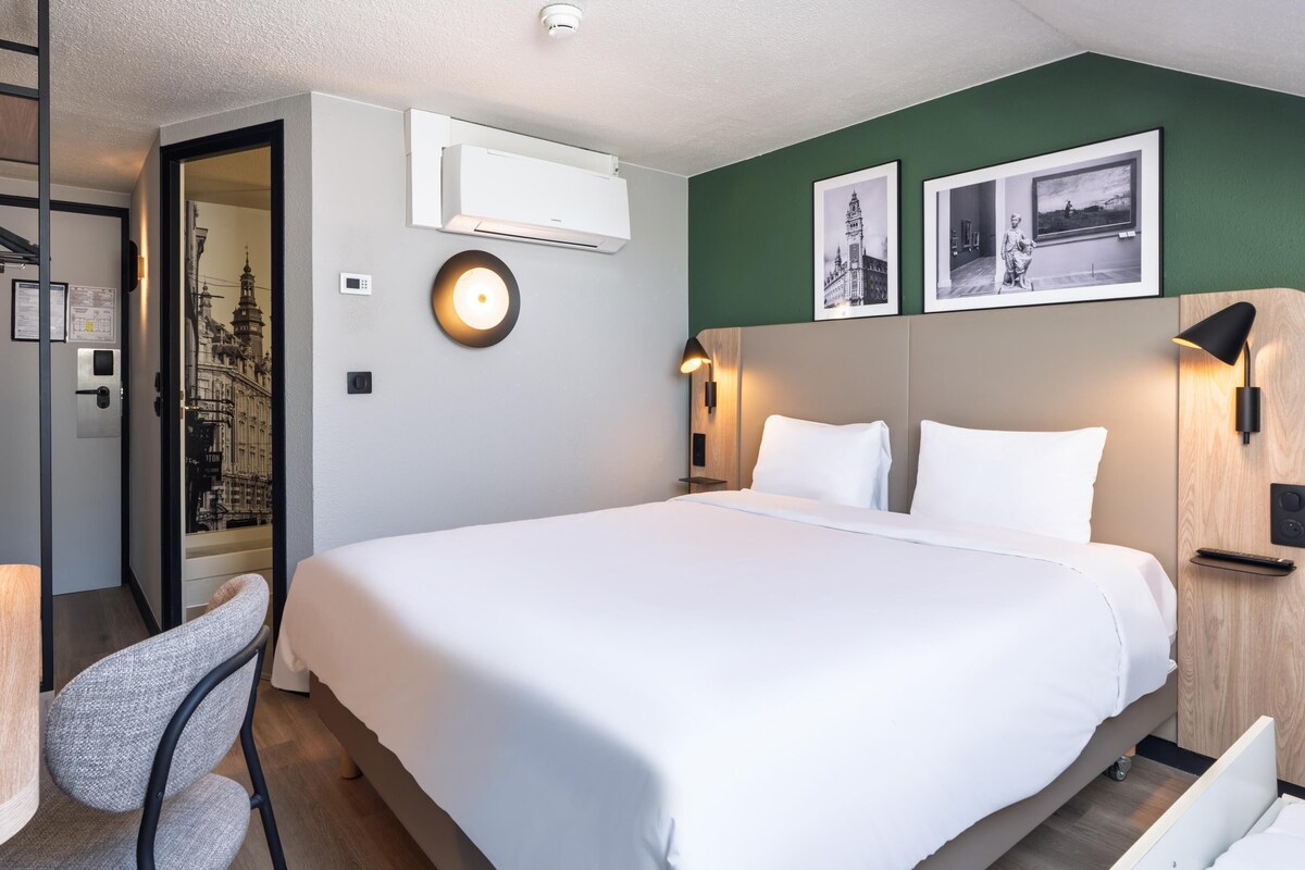 A hotel room with a bed, a desk, a chair, an air conditioning unit, and two framed pictures on the wall.