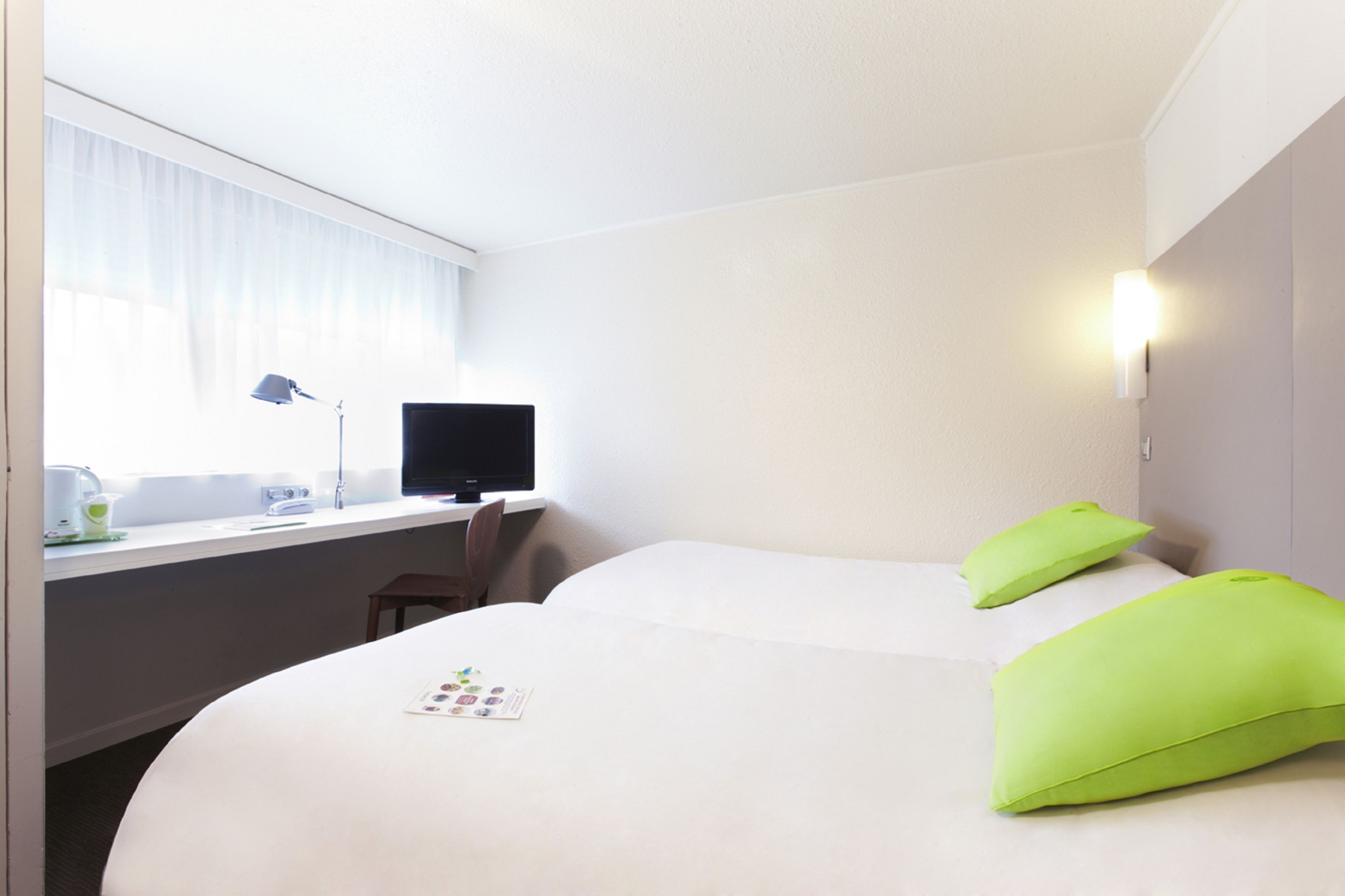 Una camera d'albergo con due letti, una scrivania con un computer, una lampada e una finestra con tende.