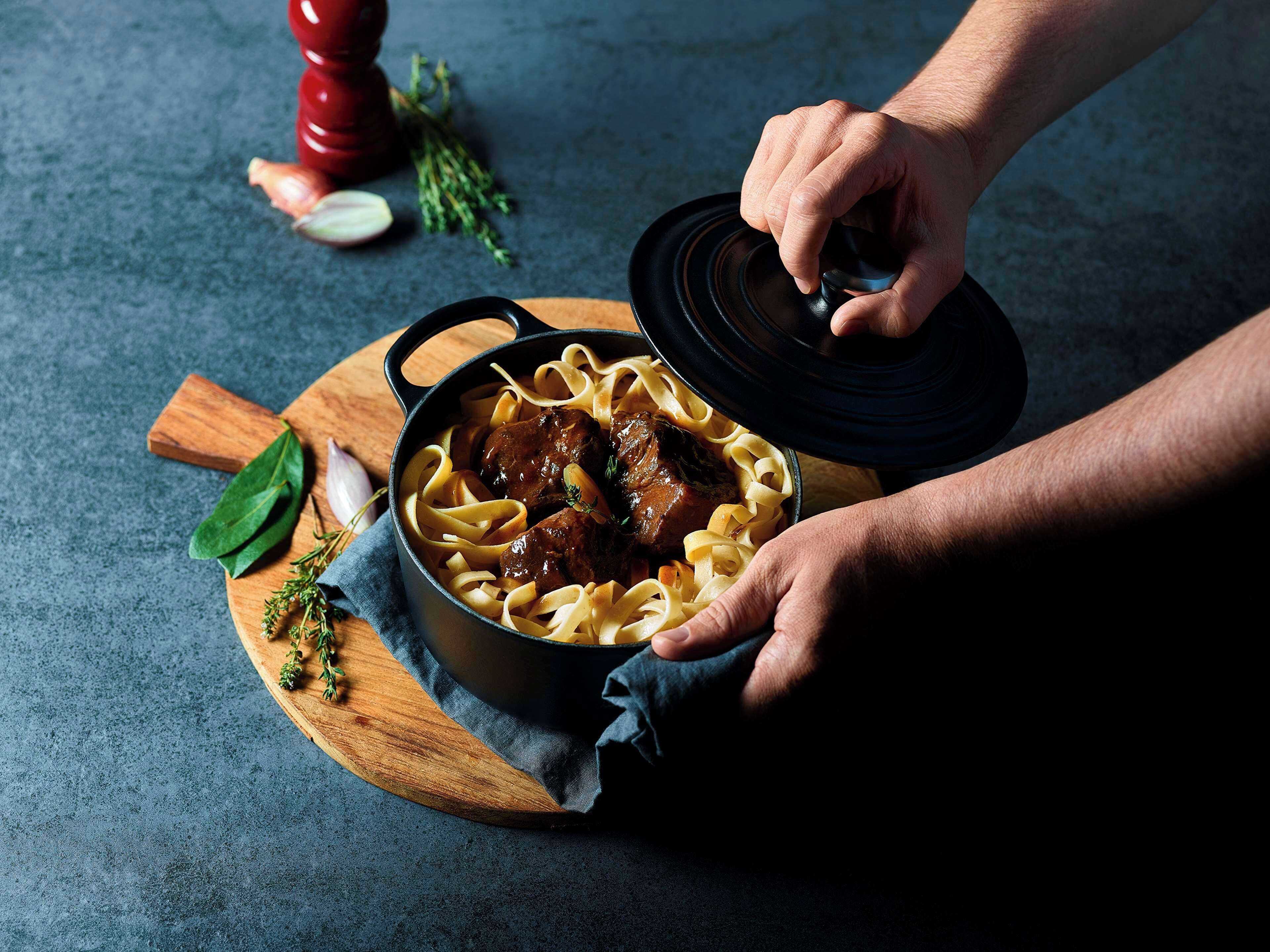 A person lifting a lid off a pot containing a stew with noodles.