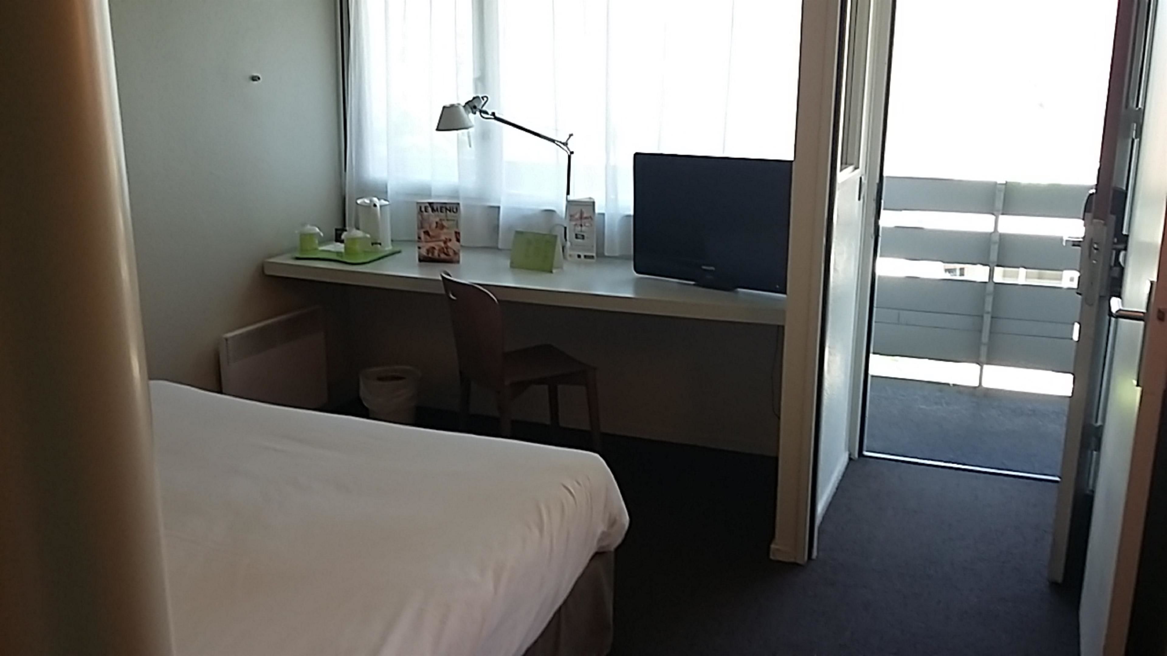 Une chambre d’hôtel avec un lit, un bureau avec un ordinateur, une chaise et une fenêtre avec des rideaux.