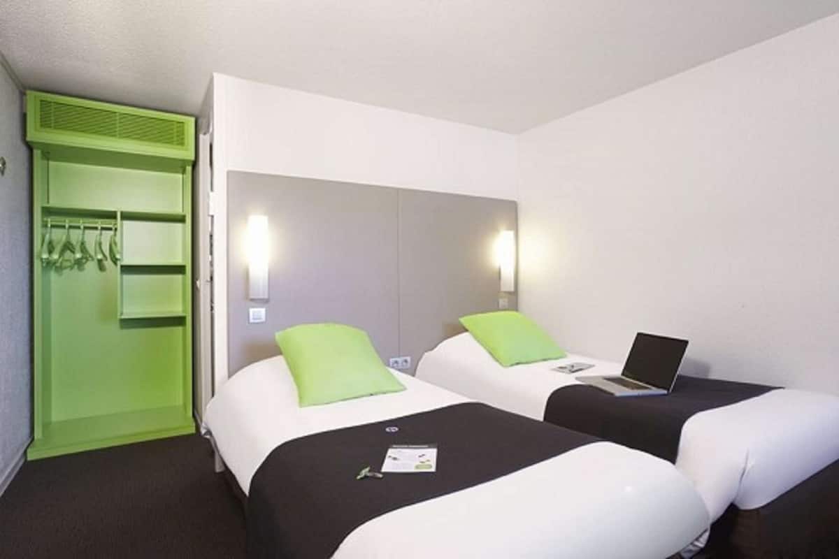 Une chambre d’hôtel avec deux lits, chacun agrémenté d’un oreiller vert et d’un ordinateur portable posé sur l’un des lits. On y trouve une armoire verte contenant des vêtements suspendus, et un miroir au-dessus des lits.