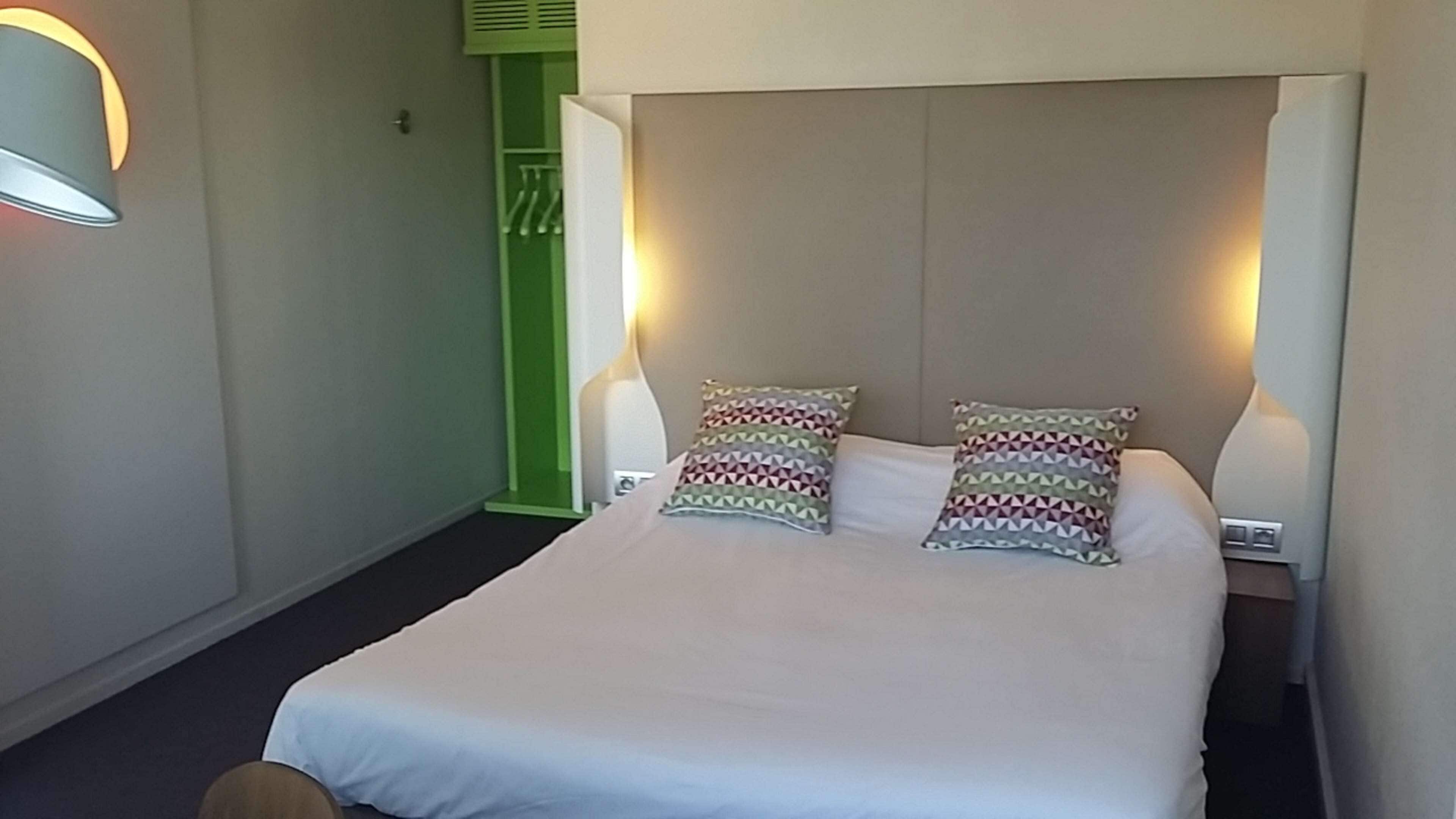 A hotel room with a large bed, two pillows, bedside lamps, a green door, and a wall-mounted light fixture.