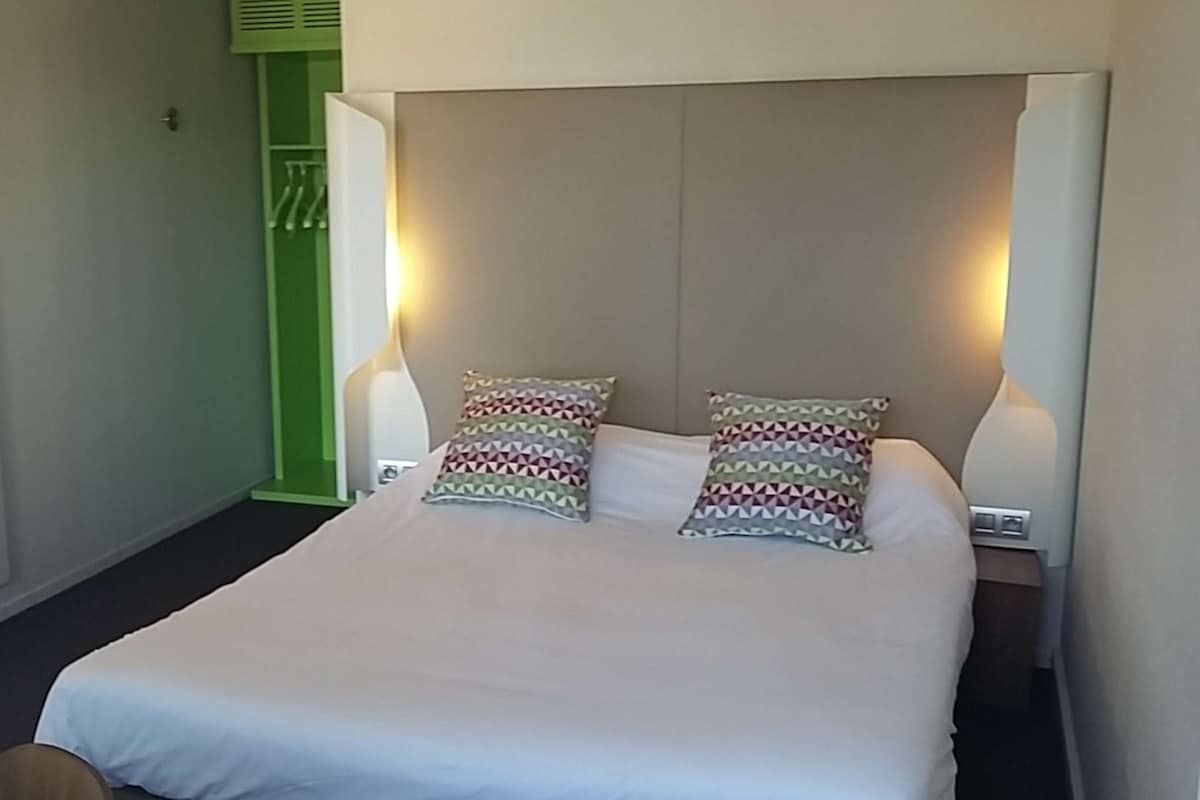 Habitación de hotel con una cama grande, dos almohadas, lámparas de noche, una puerta verde y una lámpara fijada en la pared.