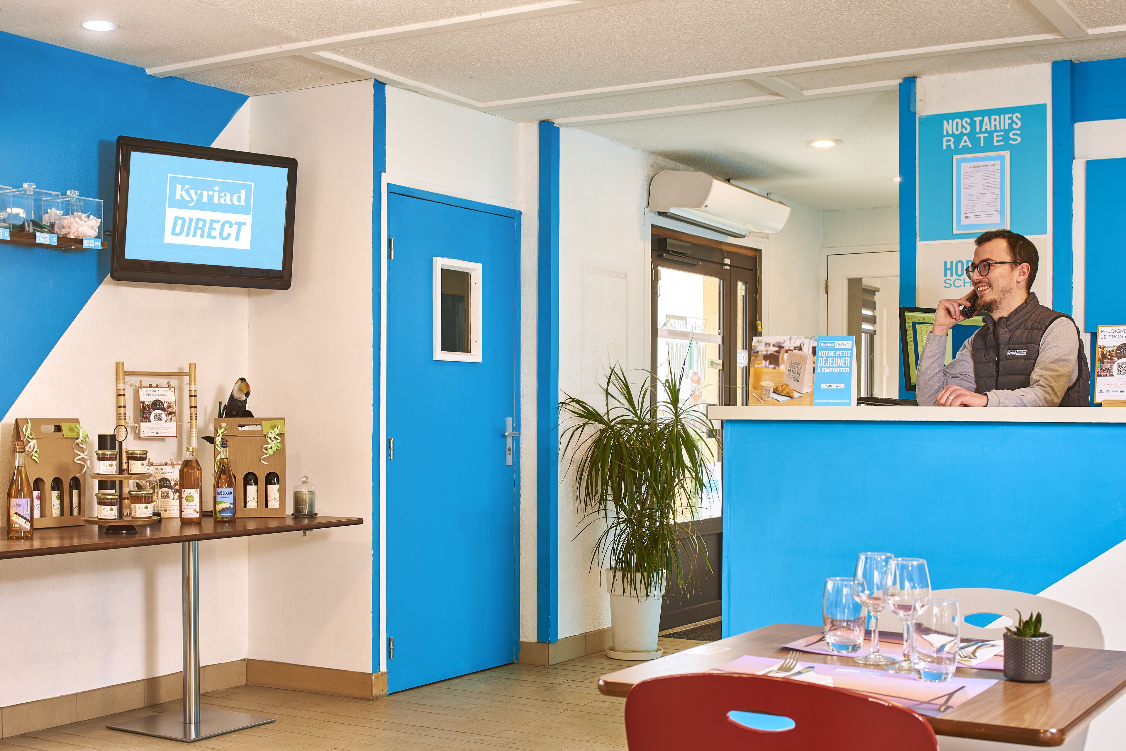 Uma área de recepção com esquema de cores azul e branco, uma tela exibindo 'Kyriad DIRECT' e um balcão com um vaso de planta e garrafas de vinho.