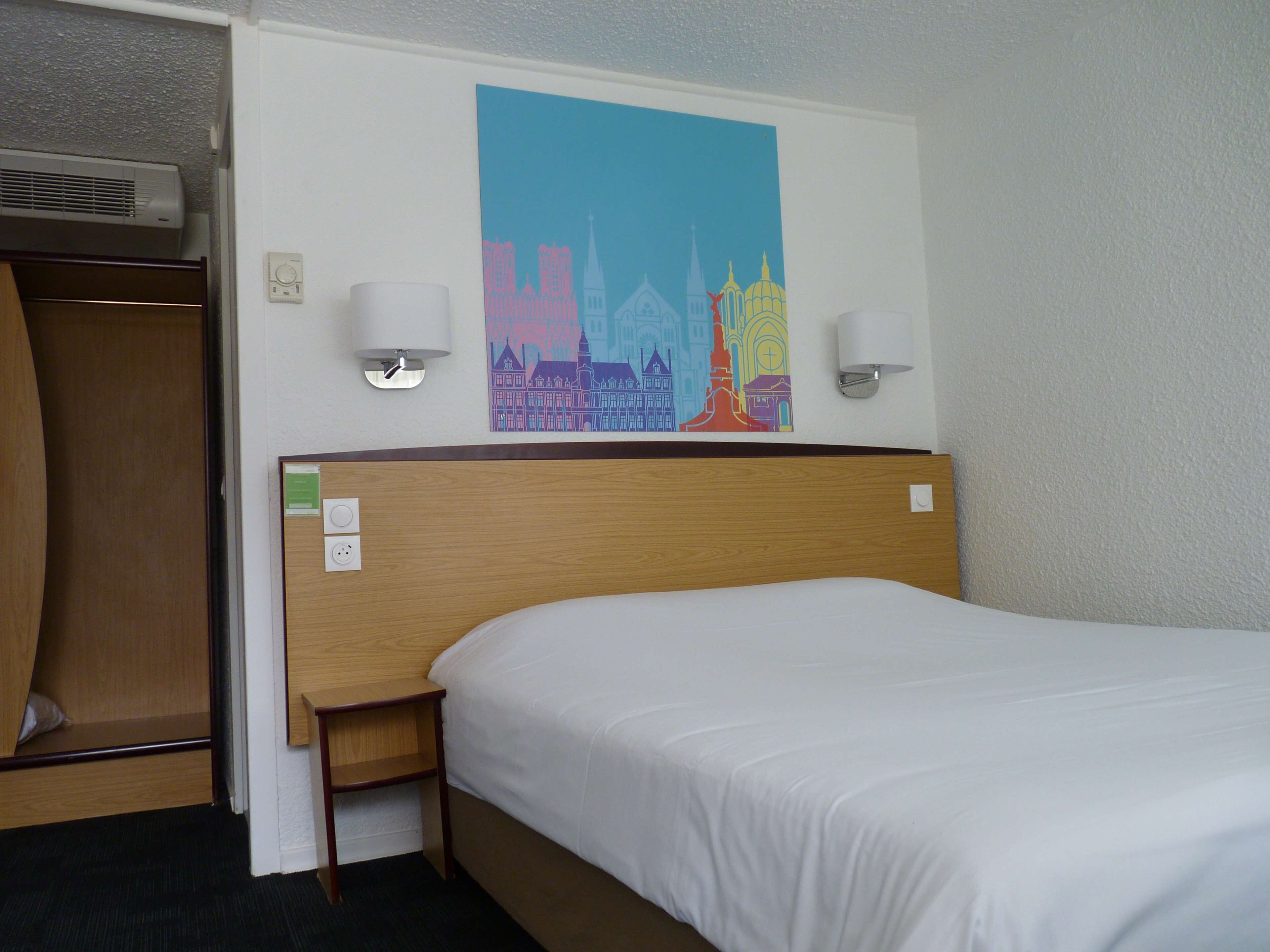 Habitación de hotel con cama, cabecera de madera, dos lámparas fijadas a la pared, aire acondicionado instalado en la pared y una obra de arte con un paisaje urbano colorido.