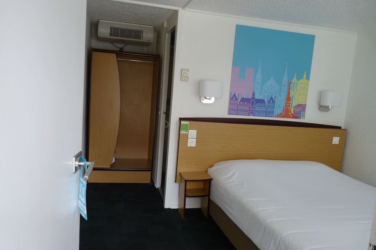 A hotel room with a bed, a wooden headboard, a nightstand, a wall-mounted lamp, a painting, and a door leading to a bathroom.