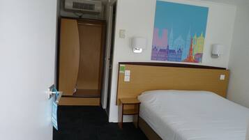 A hotel room with a bed, a wooden headboard, a nightstand, a wall-mounted lamp, a painting, and a door leading to a bathroom.