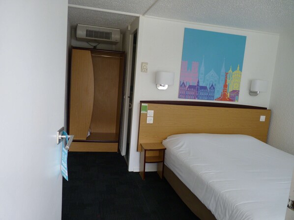 A hotel room with a bed, a wooden headboard, a nightstand, a wall-mounted lamp, a painting, and a door leading to a bathroom.