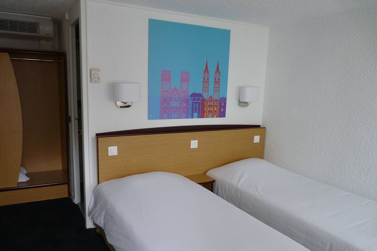 A hotel room with two single beds, a wooden headboard, a wall-mounted air conditioner, and a colorful cityscape painting above the beds.
