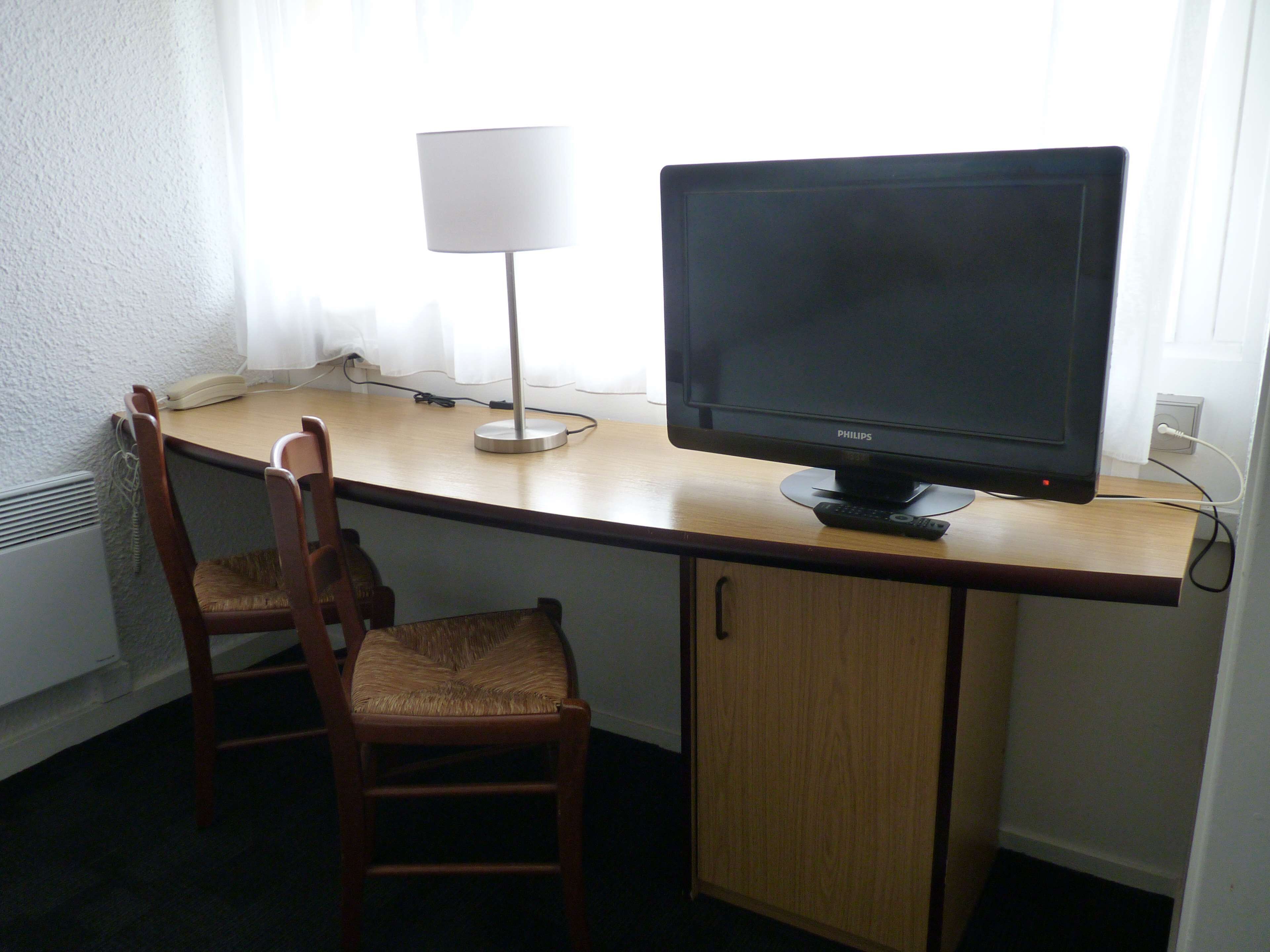 A hotel room with a wooden desk, two wooden chairs, a black monitor, a white lamp, and a white radiator.
