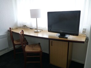 A hotel room with a wooden desk, two wooden chairs, a black monitor, a white lamp, and a white radiator. - Kyriad Direct Le Havre Est - Gonfreville (Gonfreville-l'Orcher)