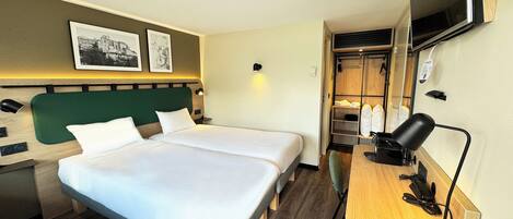 A hotel room with two beds, a desk with a computer, a TV mounted on the wall, and a closet with hanging clothes.