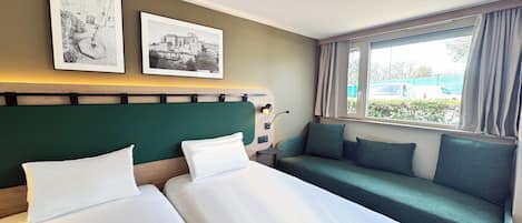 A hotel room with two beds, a green headboard, a window with a view of the outside, and two framed pictures on the wall.