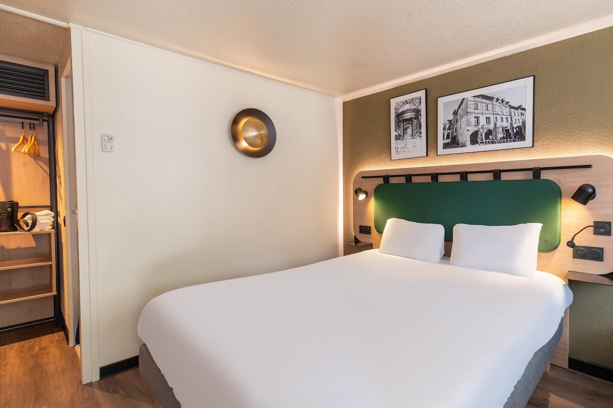 Ein ordentlich bezogenes Bett in einem Hotelzimmer, mit einem grünen Kopfteil, zwei gerahmten Bildern an der Wand und einem kleinen Nachttisch mit einer Lampe.