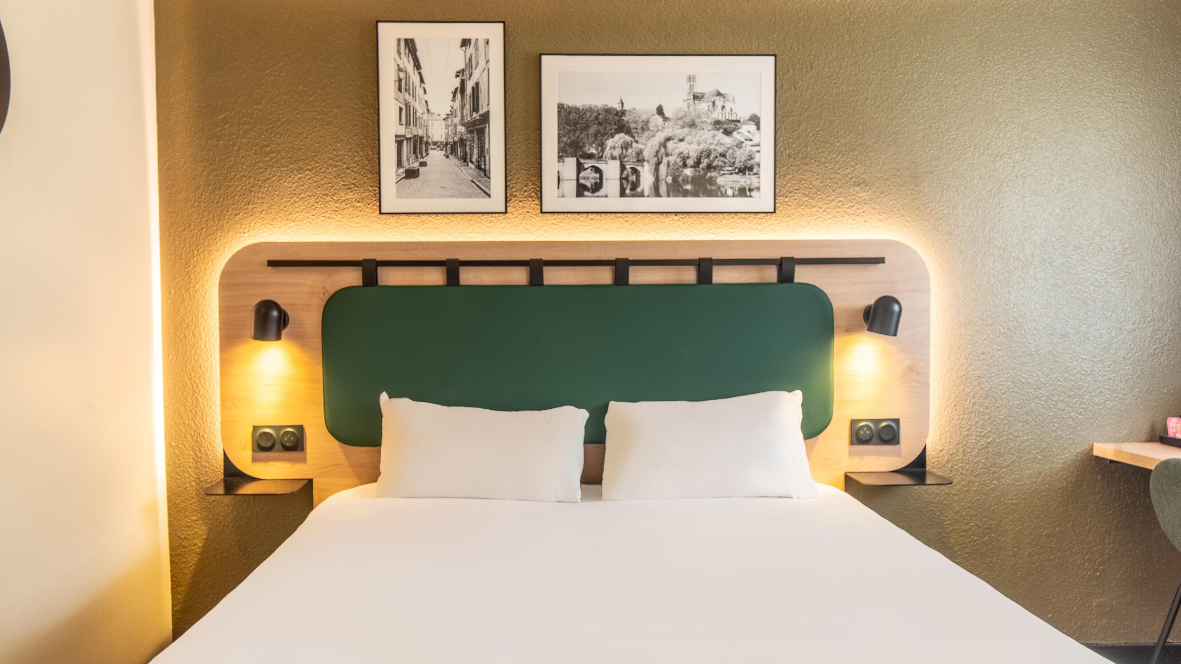 A hotel room with a green upholstered headboard, two bedside lamps, a white bedspread, and two framed black-and-white photographs on the wall.