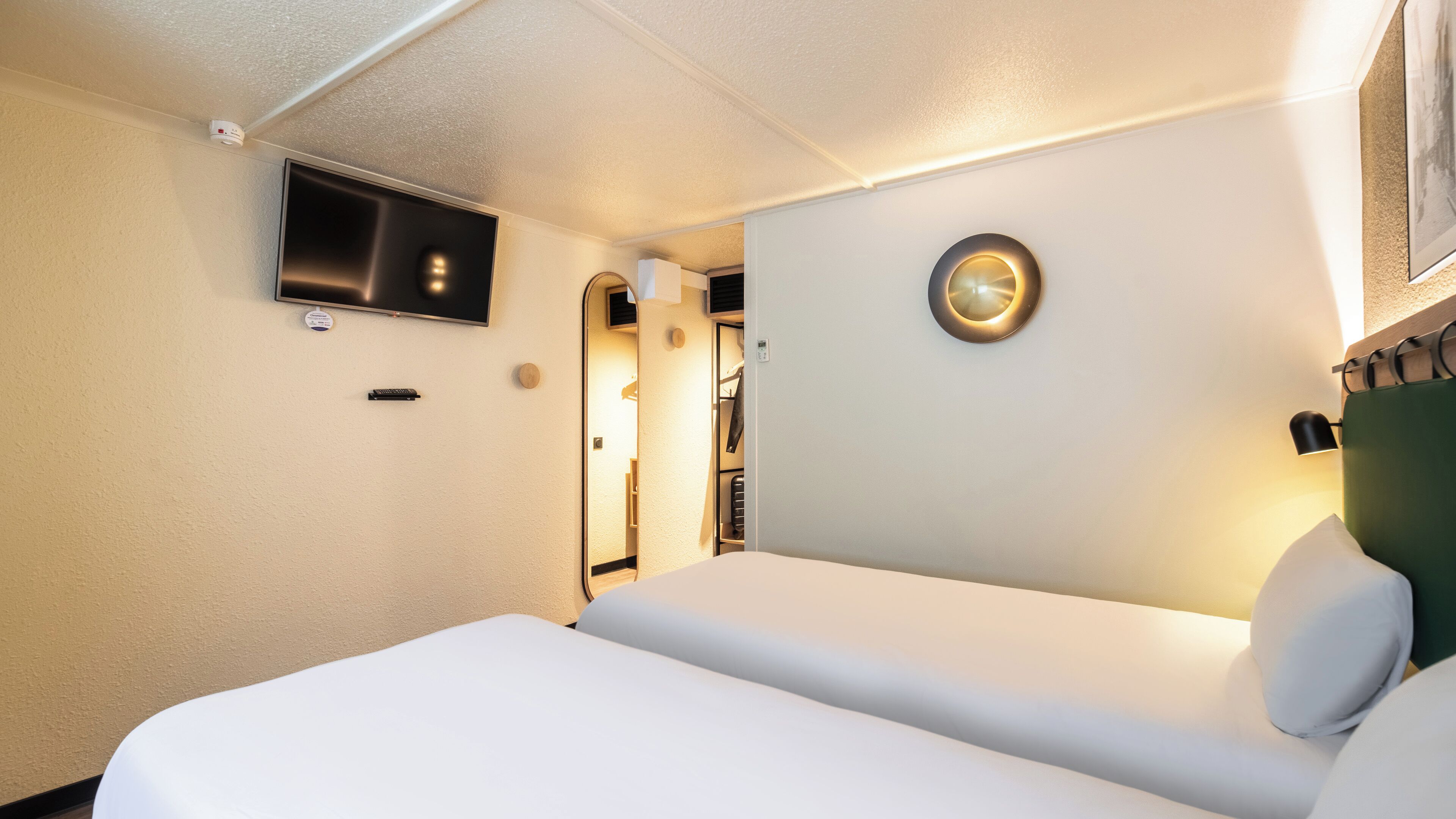 Une chambre d’hôtel avec un lit, un téléviseur fixé au mur, un applique murale ronde et un porte-manteau.