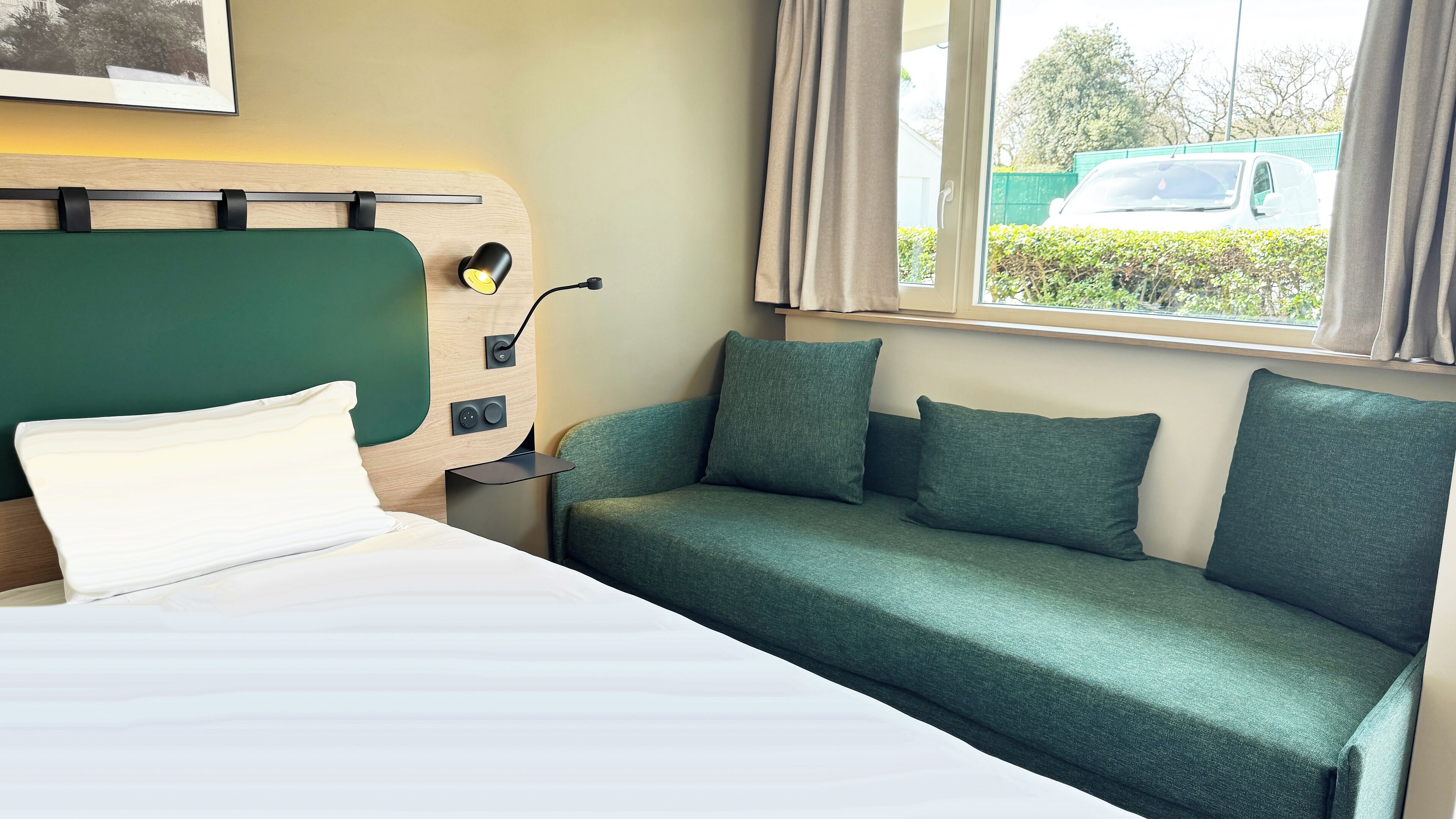 A hotel room with a bed, a green sofa, a window with curtains, and a view of a parked car outside.