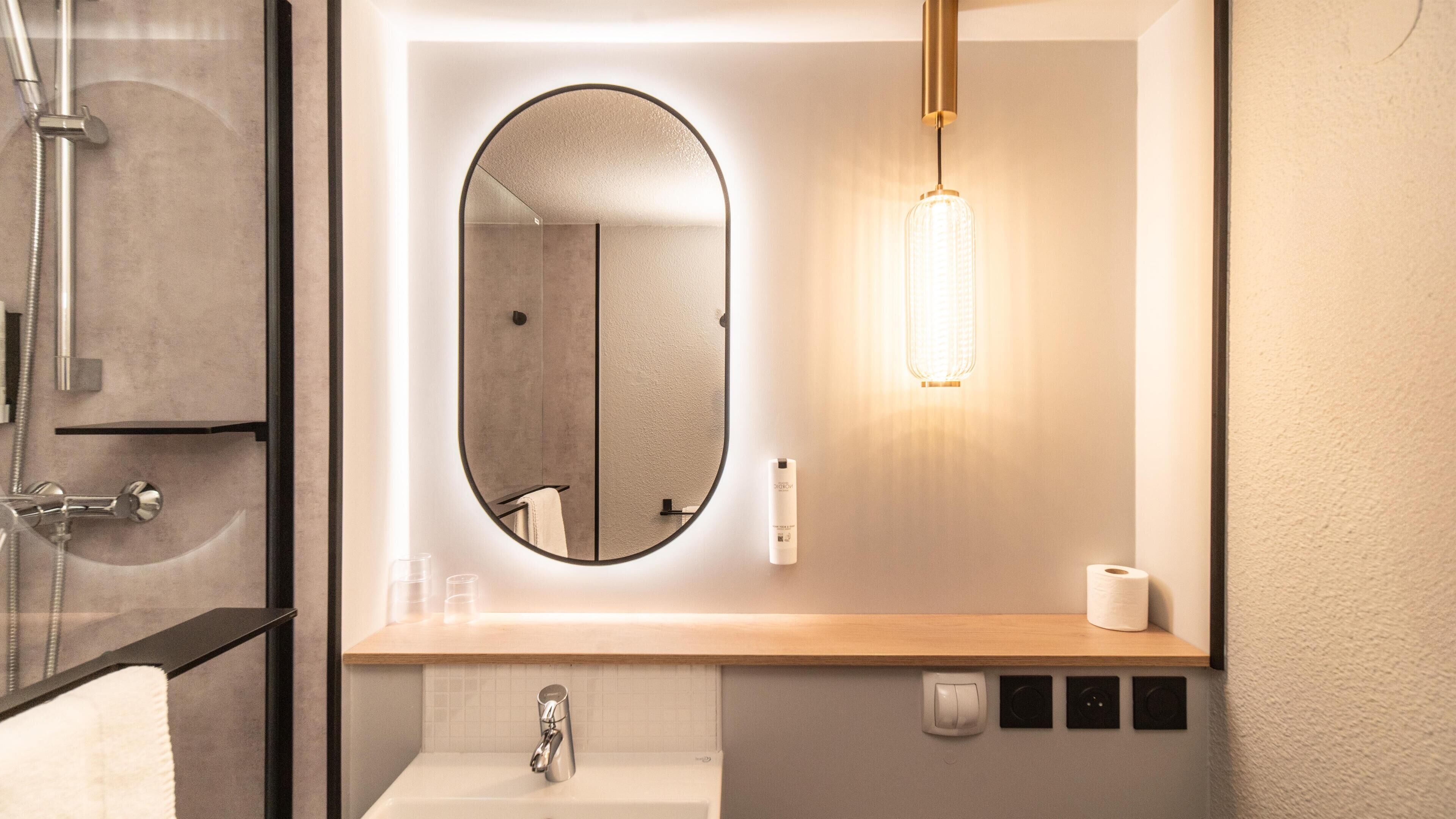 A modern bathroom with a round mirror, a wooden countertop, a sink, a handheld showerhead, and a wall-mounted light fixture.