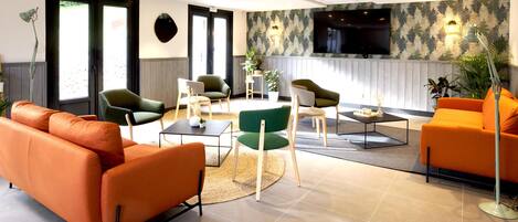 A modern hotel lobby with a seating area featuring orange sofas and green chairs, a flat-screen TV mounted on the wall, and a large window providing natural light.