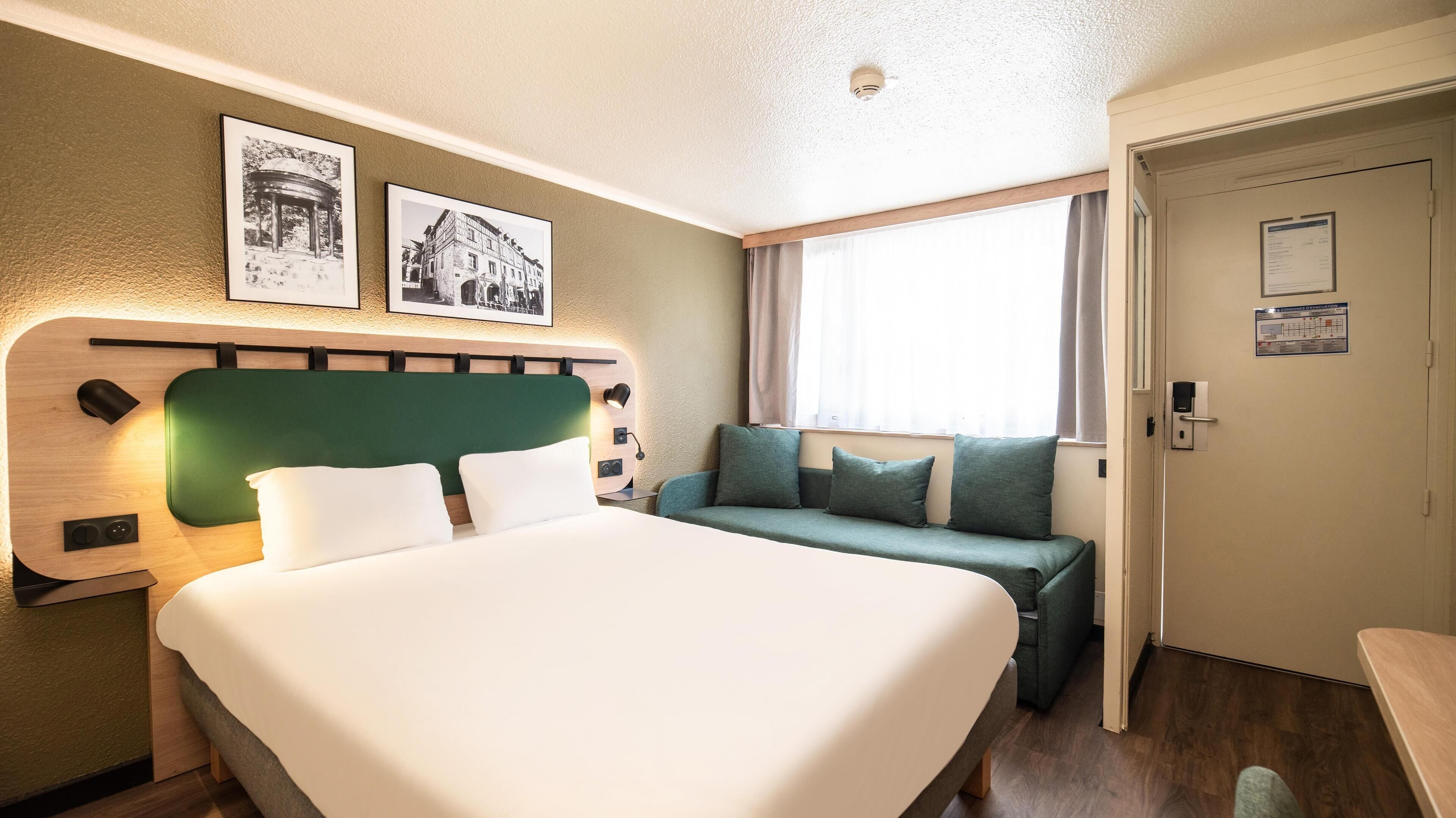 Habitación de hotel con una cama grande, un sofá verde, un cabecero de madera y dos cuadros con marcos negros en la pared.