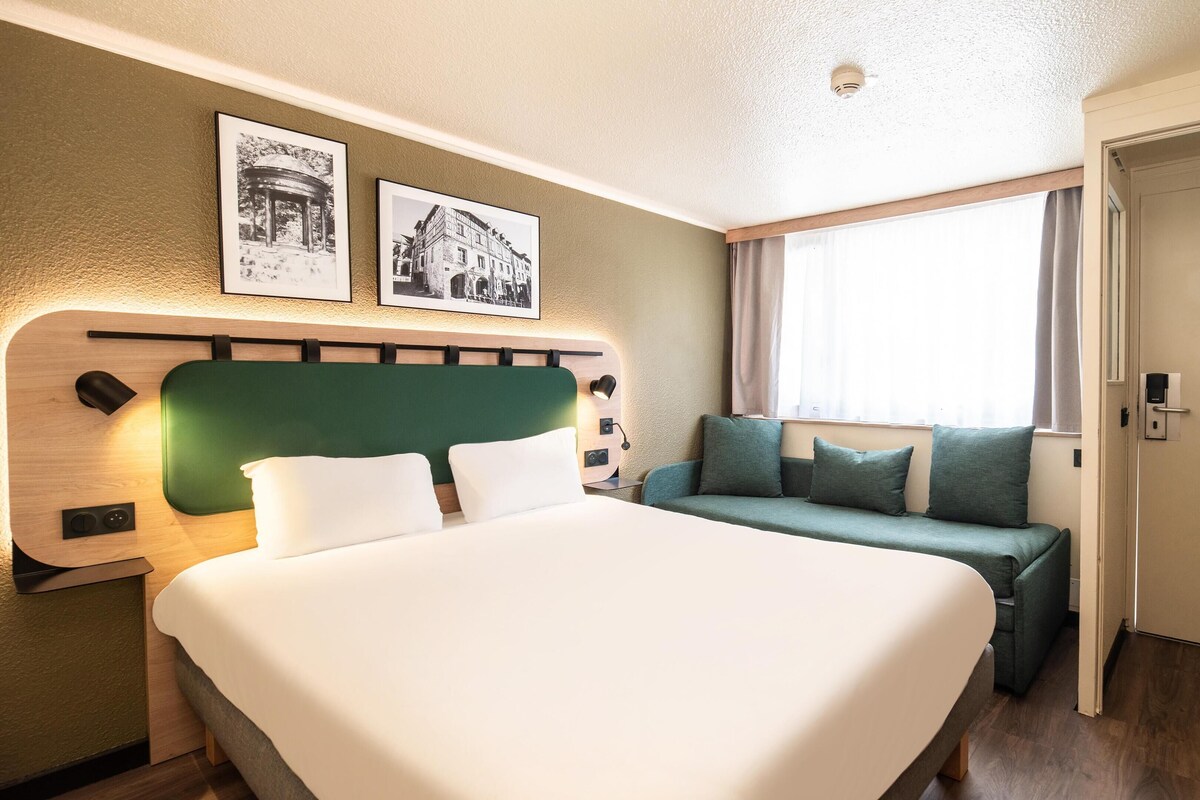 A hotel room with a large bed, a green sofa, a wooden headboard, and two black-framed pictures on the wall.