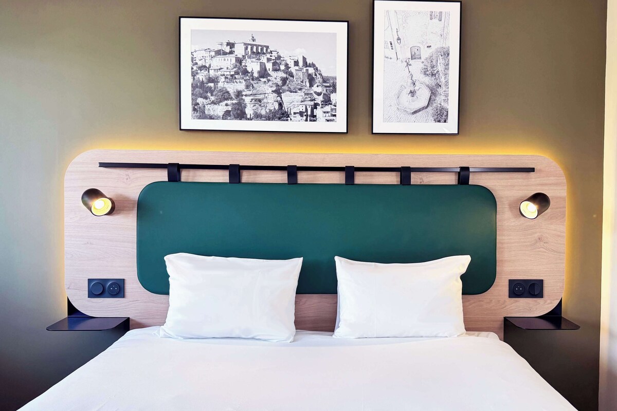 A neatly made bed with a green upholstered headboard, two white pillows, and two bedside lamps. Above the bed, there are two framed black-and-white photographs on the wall.