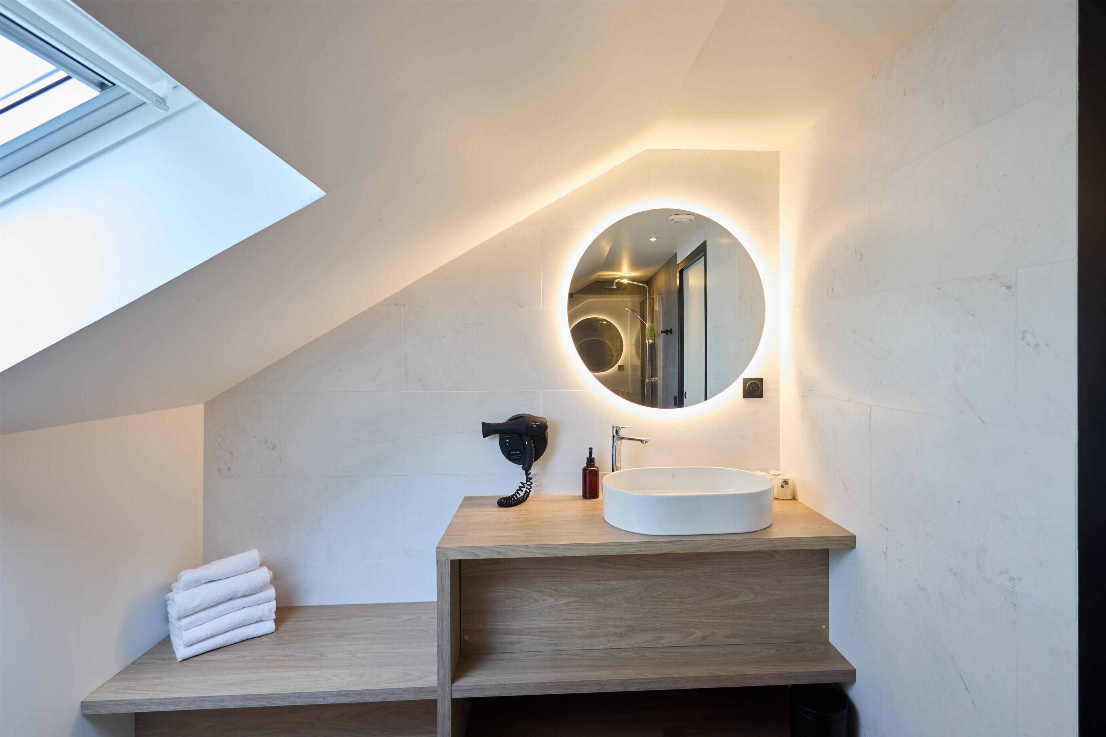 A modern bathroom with a skylight, a round mirror, a wooden vanity, and a white sink.