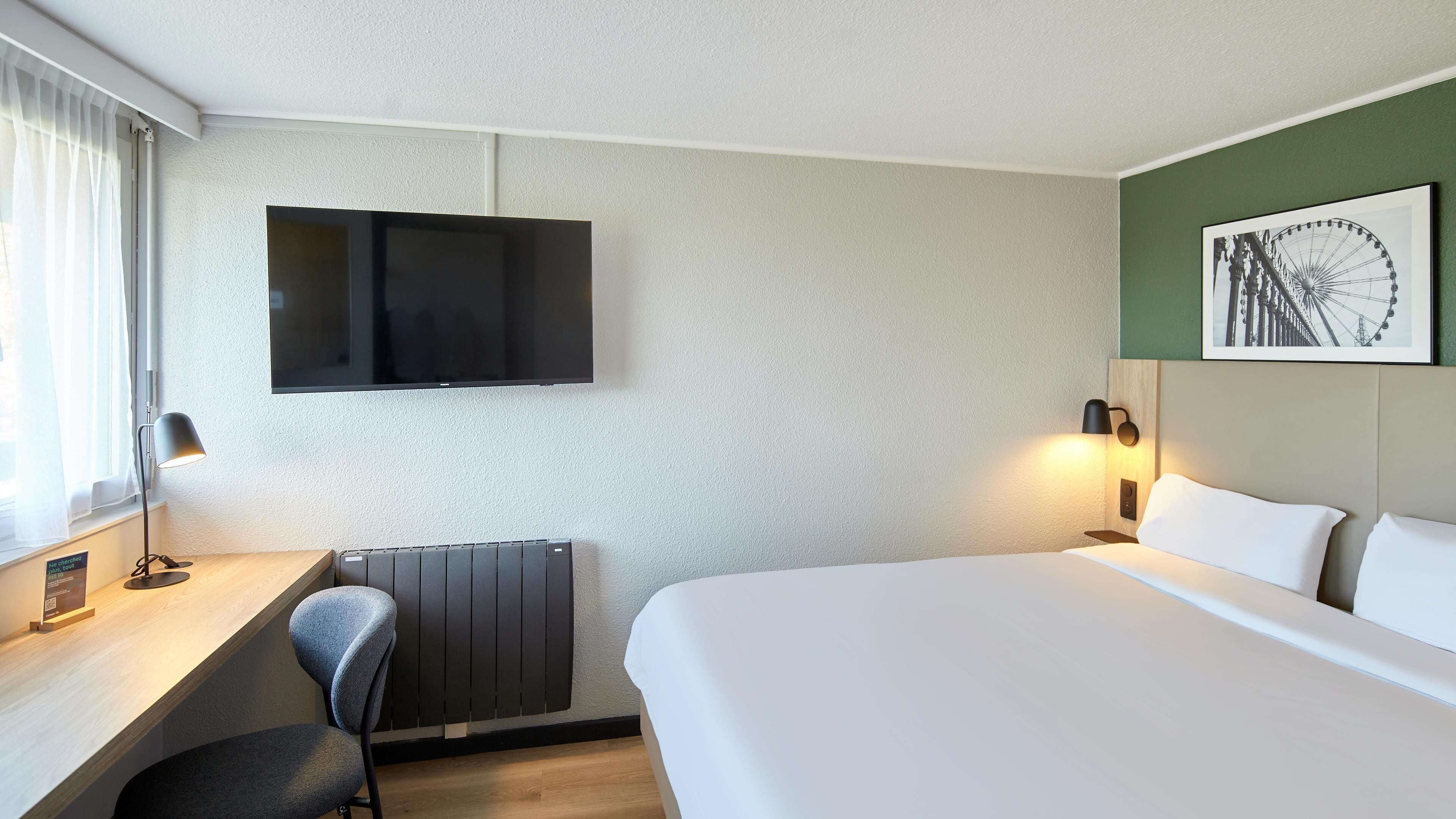 Een hotelkamer met een bed, een bureau met stoel, een aan de muur bevestigde televisie en een afbeelding van een reuzenrad.