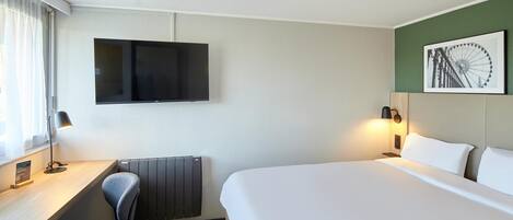 A hotel room with a bed, a desk with a chair, a TV mounted on the wall, and a picture of a Ferris wheel.