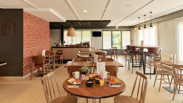 Een restaurant met houten tafels en stoelen, een bakstenen muur en een zichtbare keuken in de achtergrond.