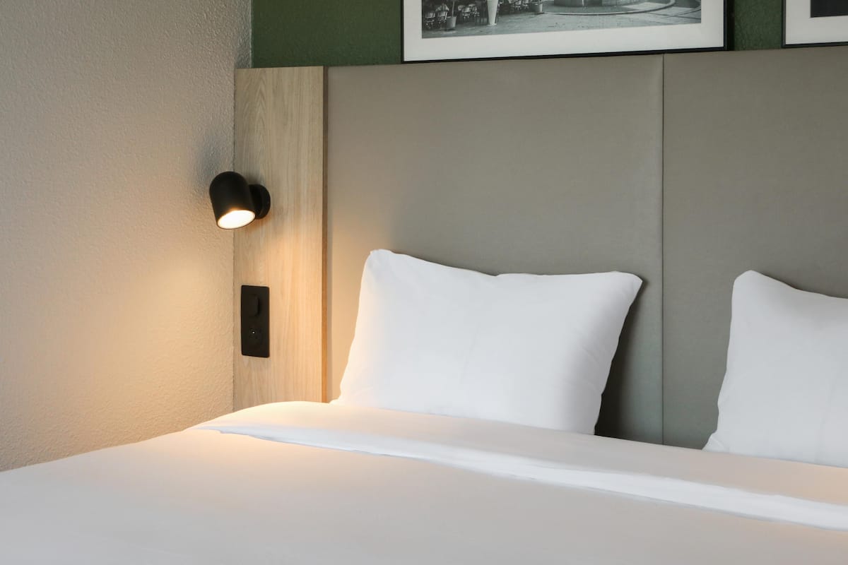 A neatly made bed with white linens, a wall-mounted light fixture, and a framed picture above the bed.