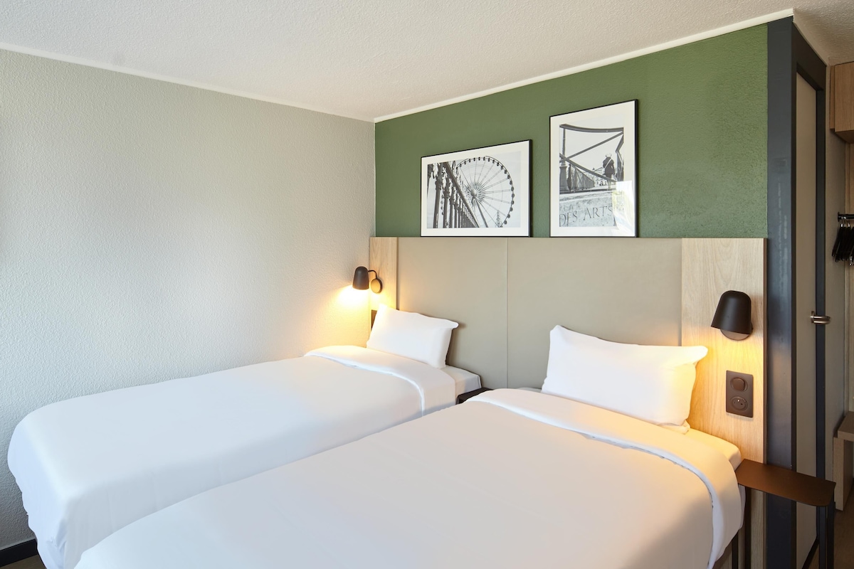 Une chambre d’hôtel avec deux lits, un téléviseur à écran plat fixé au mur et deux tableaux encadrés au-dessus des lits.
