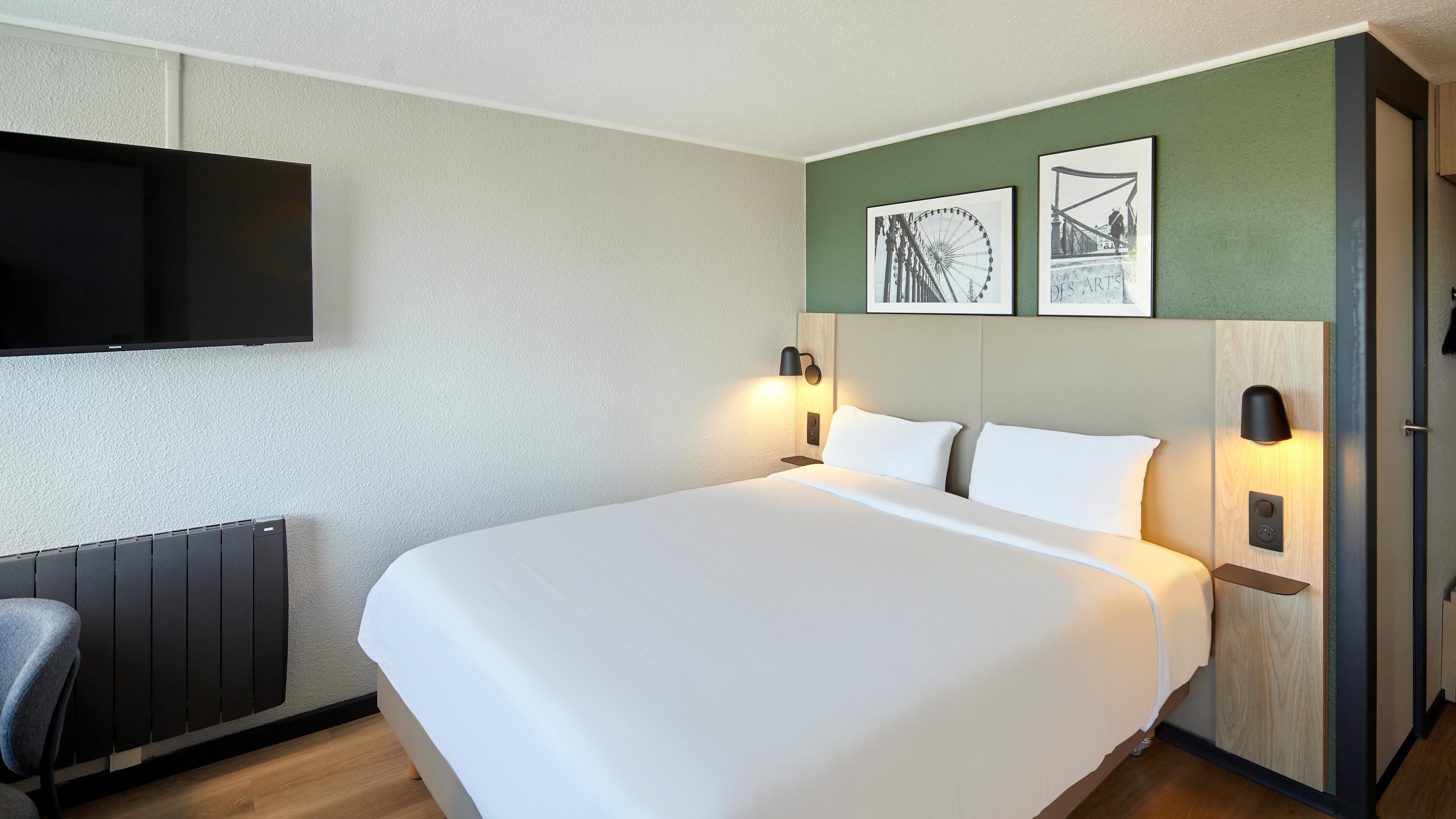 A hotel room with a large bed, two bedside tables with lamps, a flat-screen TV mounted on the wall, and two framed pictures above the bed.