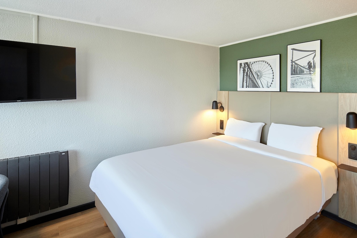 Une chambre d’hôtel avec un grand lit, deux tables de chevet avec des lampes, un téléviseur à écran plat fixé au mur et deux tableaux encadrés au-dessus du lit.