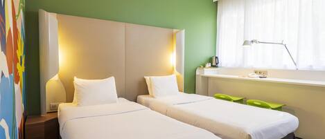 Una habitación de hotel con dos camas individuales, un escritorio con lámpara, una ventana con cortinas y una silla verde.