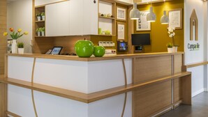 Un mostrador de recepción con acabado de madera, una pantalla digital y un reloj. Hay plantas en macetas y un certificado enmarcado en la pared.