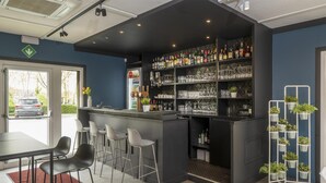 Zona de bar con mostrador, taburetes, estanterías llenas de botellas y vasos, y una decoración de plantas en macetas.