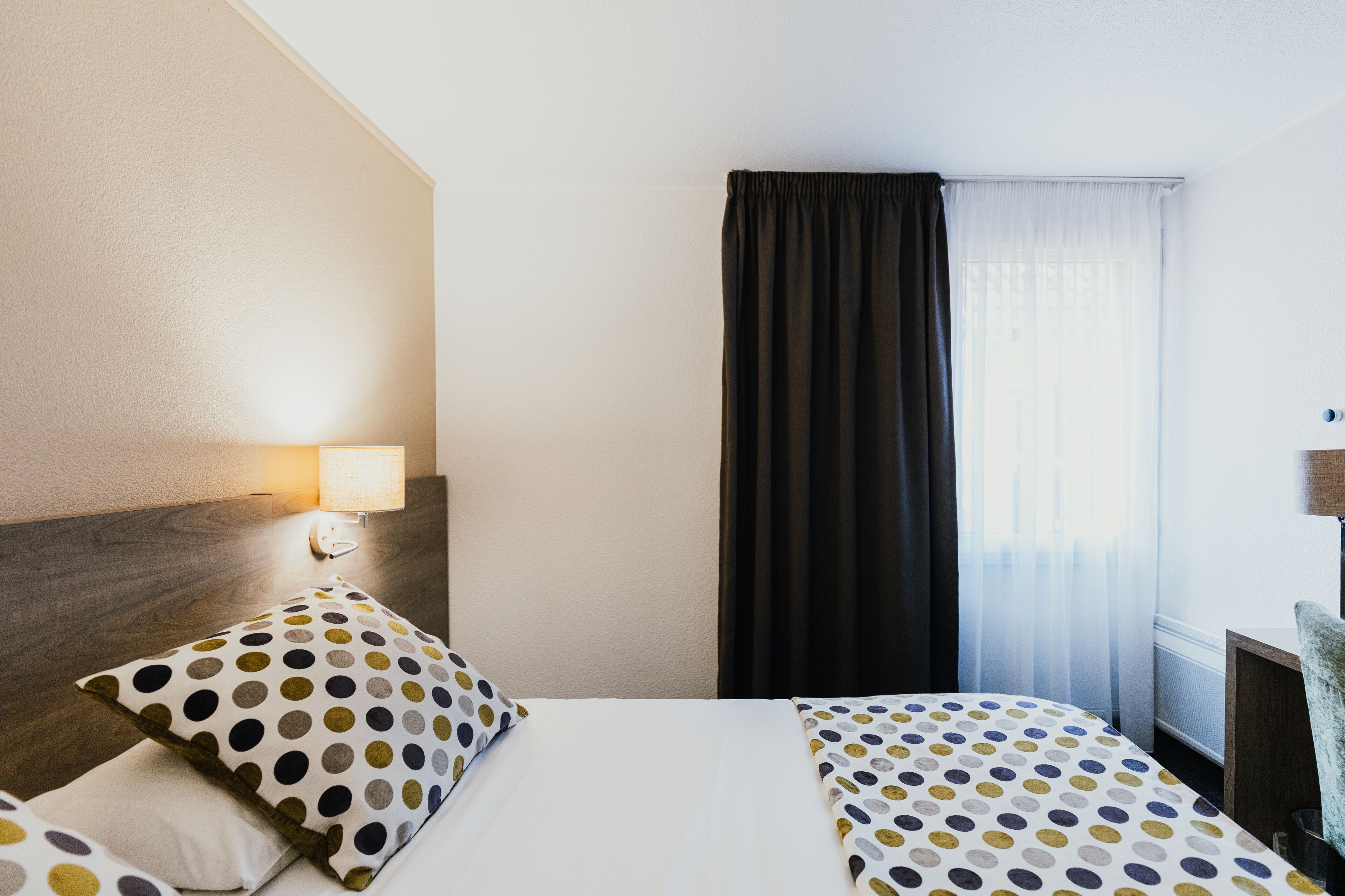 Een hotelkamer met een bed, twee kussens, een nachtkastje, een lamp en een raam met gordijnen.