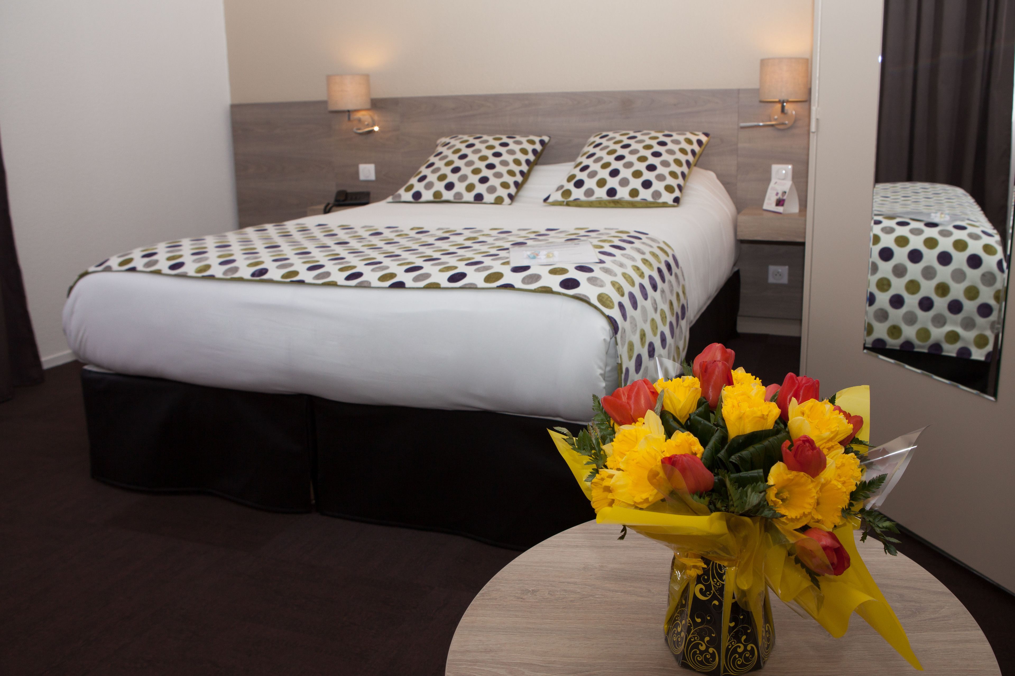 Una habitación de hotel con cama, lámparas de noche, un espejo y un jarrón con flores.