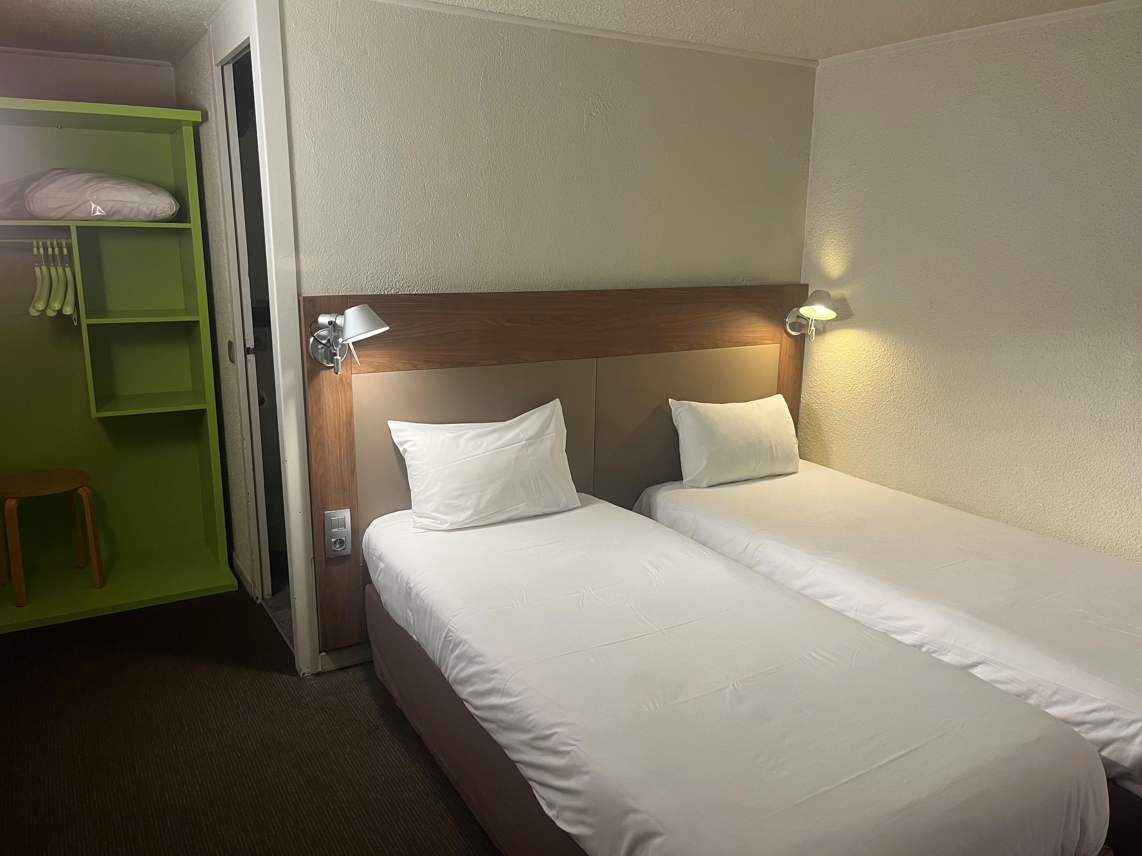 A hotel room with two single beds, a green wardrobe, and a small stool.