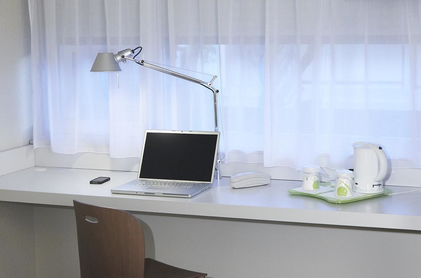 Una habitación de hotel con un escritorio blanco, una computadora portátil, una lámpara de escritorio, un teléfono, una cafetera y una mesita con tazas y un vaso.