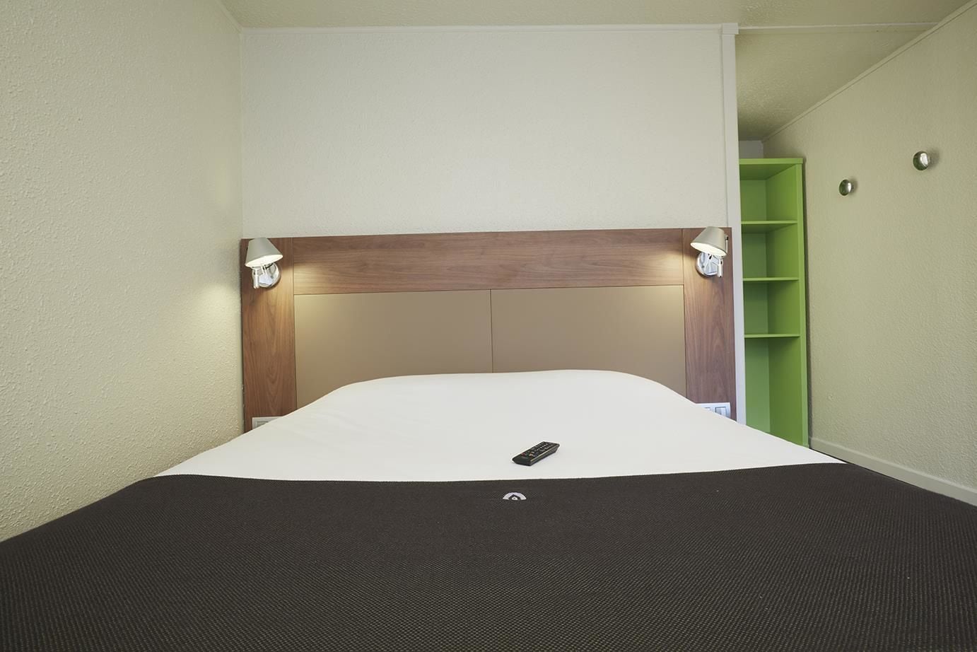 A hotel room with a large bed, a wooden headboard, two wall-mounted lamps, and a green shelving unit.